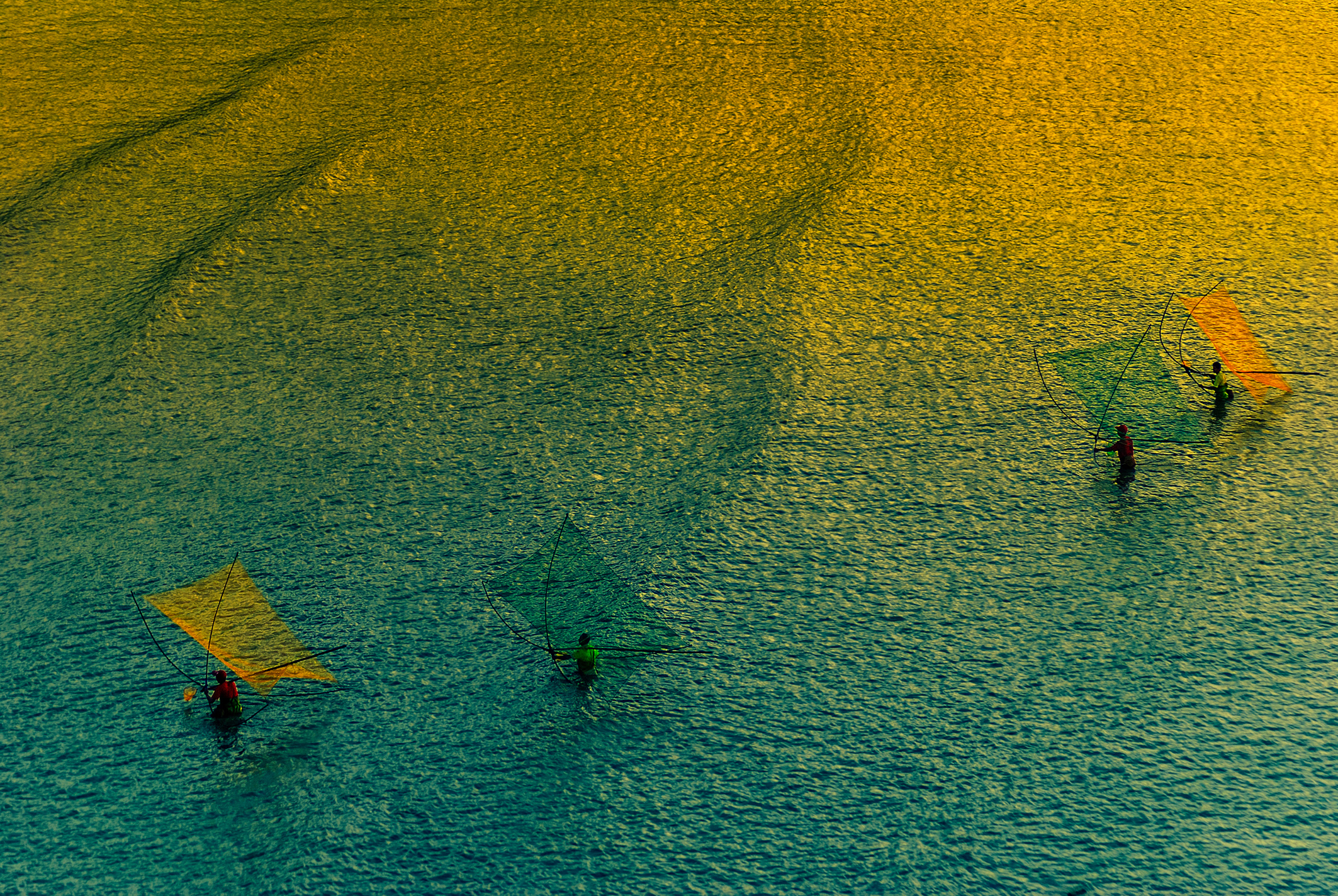 Aerial view of windsurfers gliding across teal water, with bright orange sails and dark, textured ripples creating a calm, vibrant scene