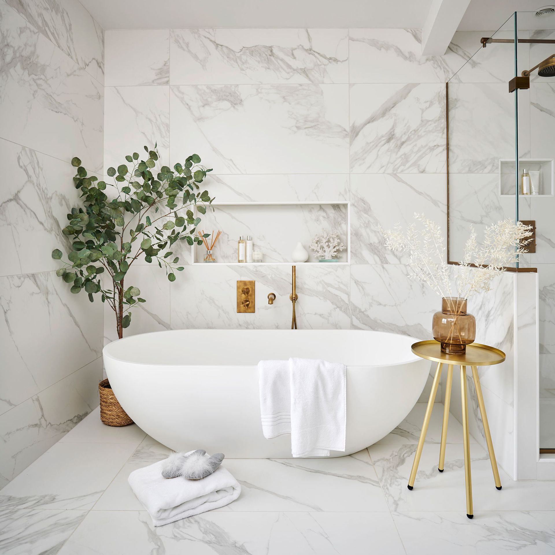 Free standing bath in front of marble wall with gold side table and large house tree in corner