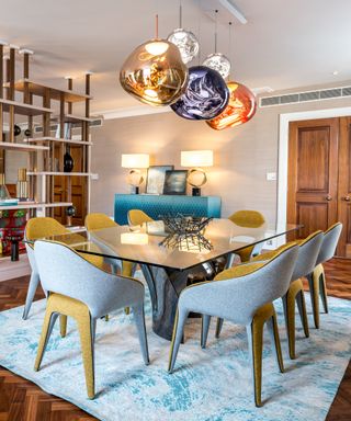 Modern dining room ideas with glass topped table, curved upholstered blue and yellow chairs, molten-like globe pendant lights and open bookshelf room divider