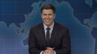 Colin Jost smiling in dark gray suit with his hands clasped around a pencil, sitting at the Weekend Update desk on Saturday Night Live Season 52