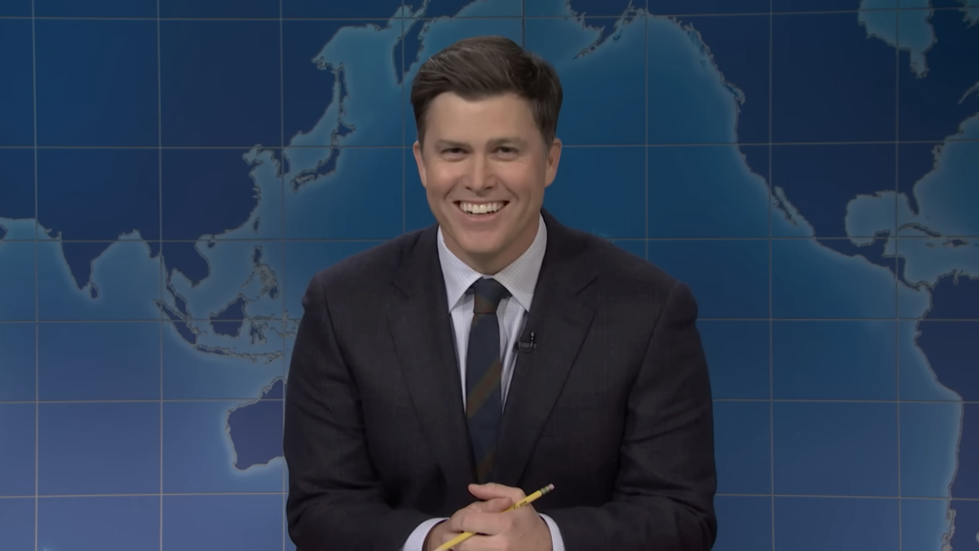 Colin Jost smiling in dark gray suit with his hands clasped around a pencil,  sitting at the Weekend Update desk on Saturday Night Live Season 52