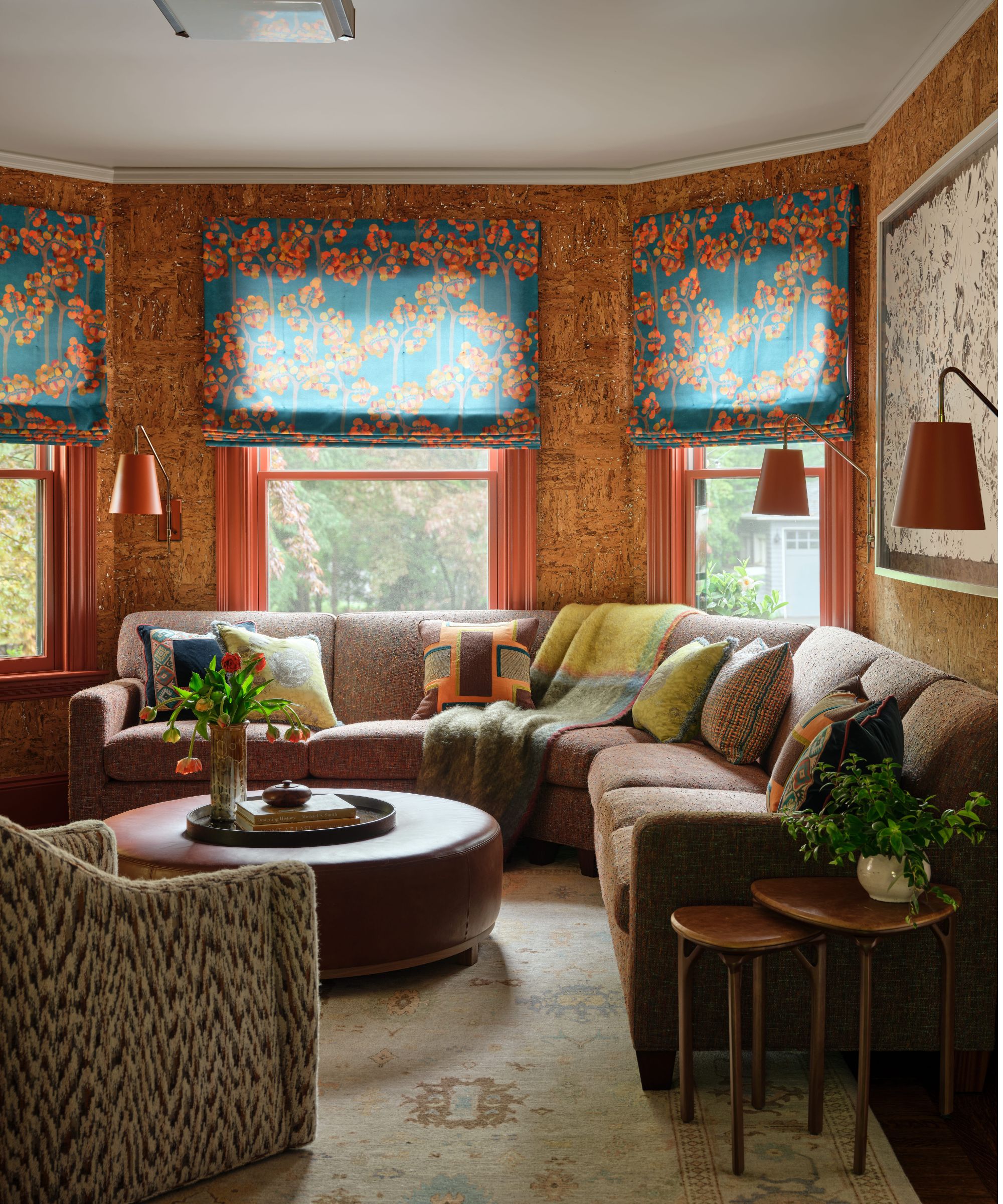 A colorful living room with a large bay window. Cork wallpaper, three red window frames with teal-and-red floral Roman blinds, a large taupe sectional with green cushions, a neutral rug, a round ottoman, and an ikat armchair.