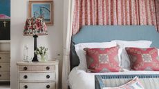 Pink pillows on a bed with a blue frame against a pink curtain; to the left, a lamp on a white nightstand against a white wall.