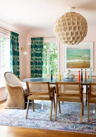 colorful dining room with pale pink walls, antique rug, wicker and wood chairs, green patterned drapes, large paper lampshade, colored candlesticks, artwork