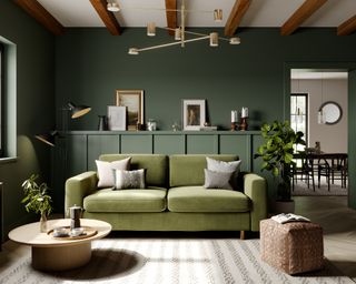 Heritage green living room scheme with deep green walls, tonal green sofa, plants, and white ceiling with exposed wooden beams.