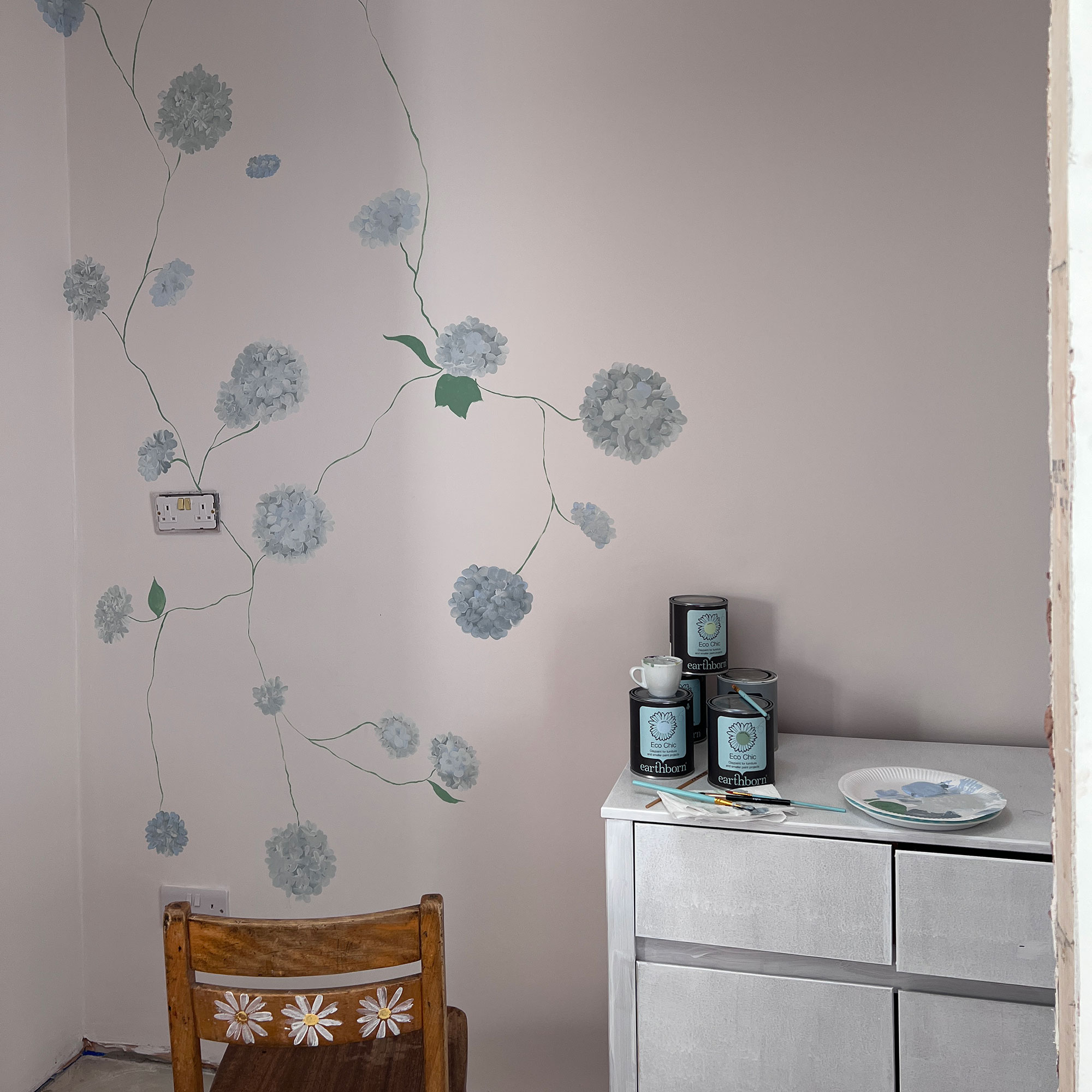 White walls being painted with blue hydrangeas, beside primed cabinet and wooden chair with three daisies painted on back