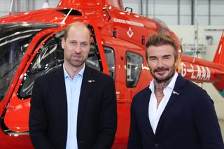 Prince William and David Beckham wearing sport jackets and smiling standing in front of a helicopter