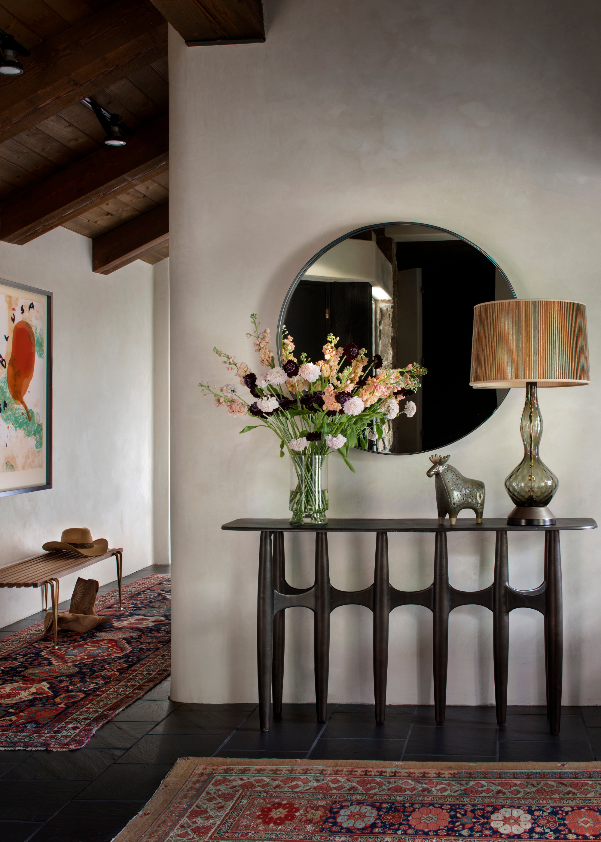 An entryway with a colorful rug and runner, by a wood side table with a vase of flowers, a lamp, and an animal statue below a round mirror