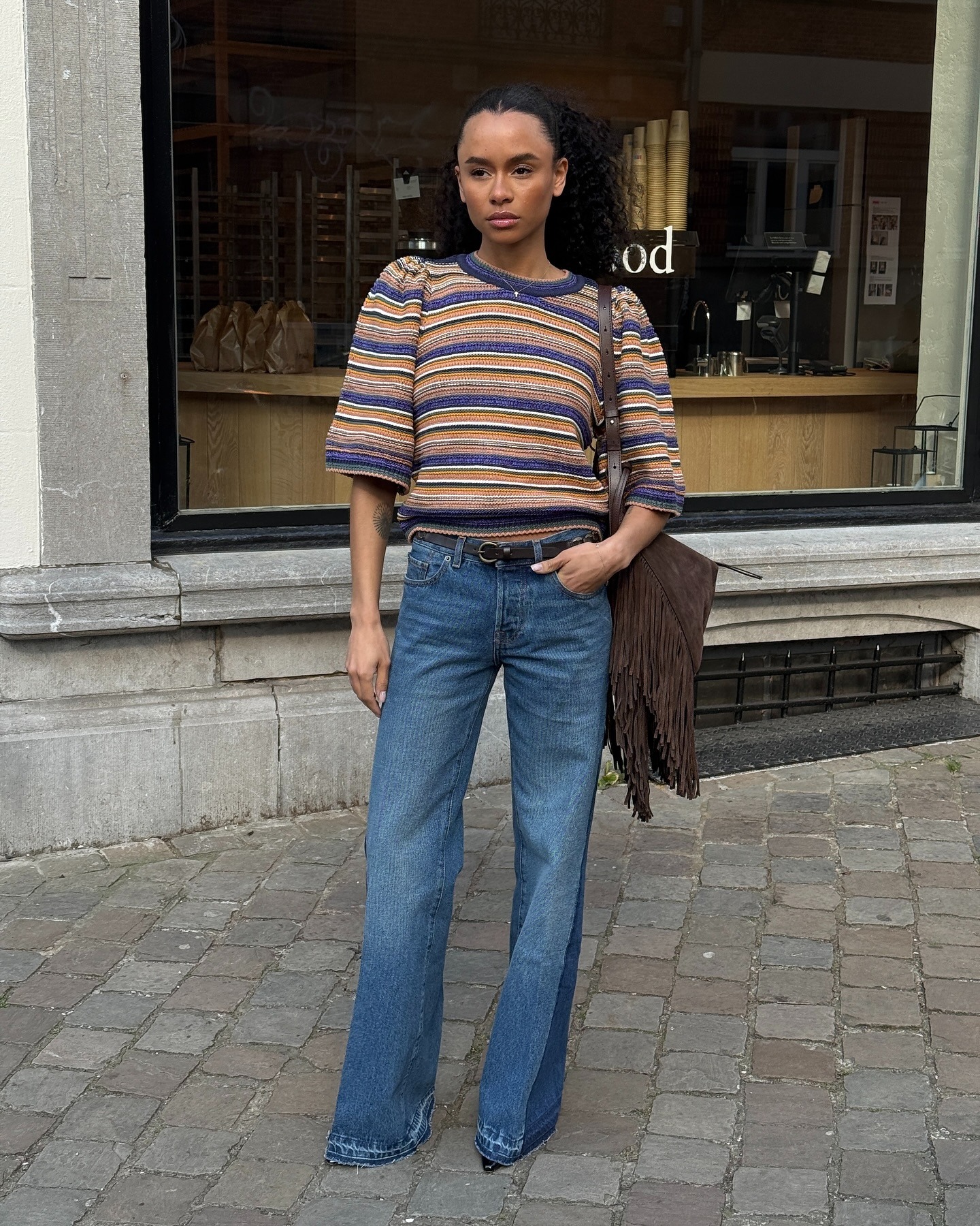 Zoe Foster wears a striped top, blue denim jean and brown suede bag. She is stood in front of a glass window in a recent image from her Instagram.