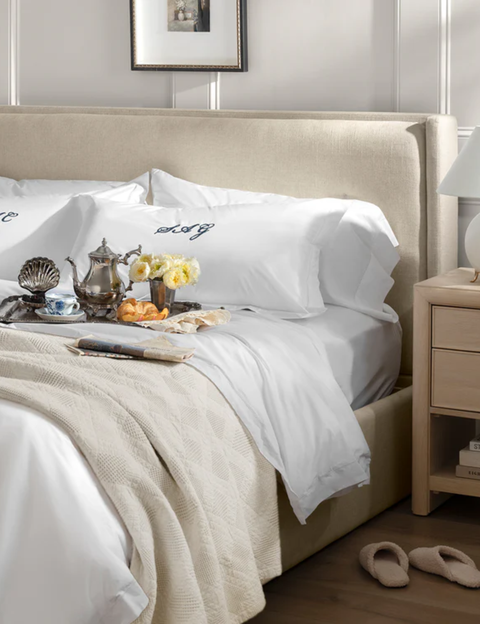 Swanky neutral bedscape featuring a pair of monogramed pillow shams and an ornate silver tray scattered with vintage-inspired silver serveware.