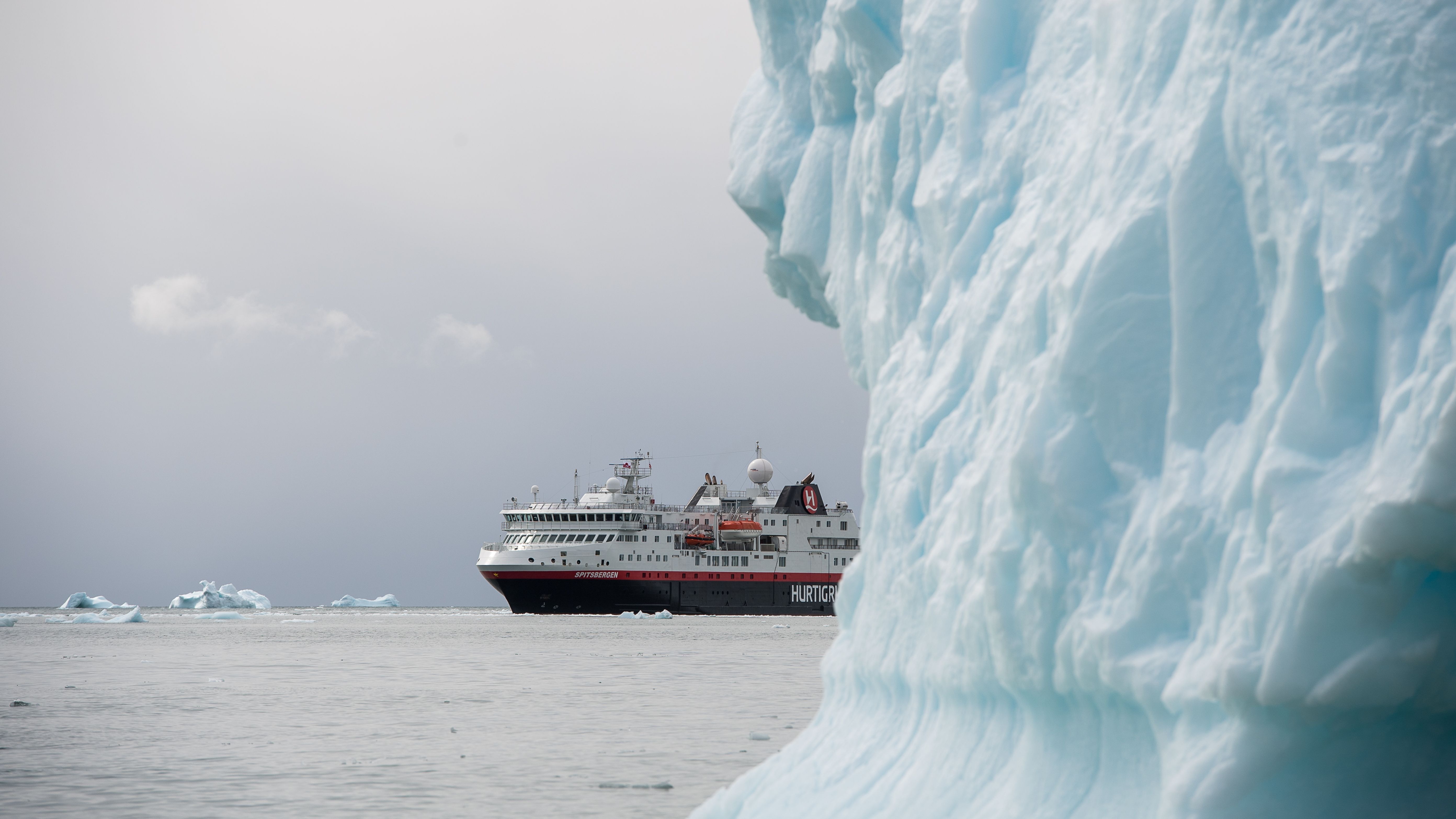 a hurtigruten ship peeks out from behind a large iceberg.