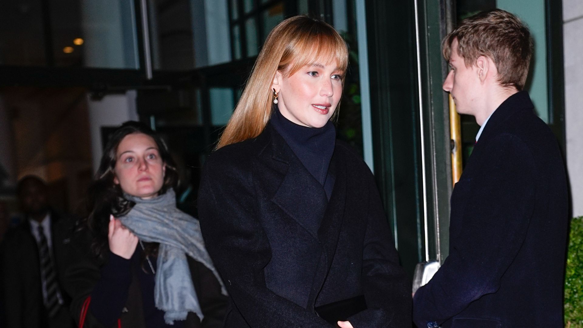 Jennifer Lawrence steps outside wearing a black coat with navy cigarette trousers with a black coat and black asymmetric heels.