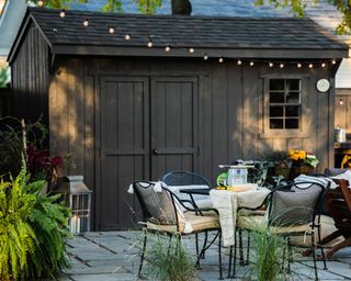 Dark hued shed with festoon lighting, and casual dining set-up on paved stone terrace.
