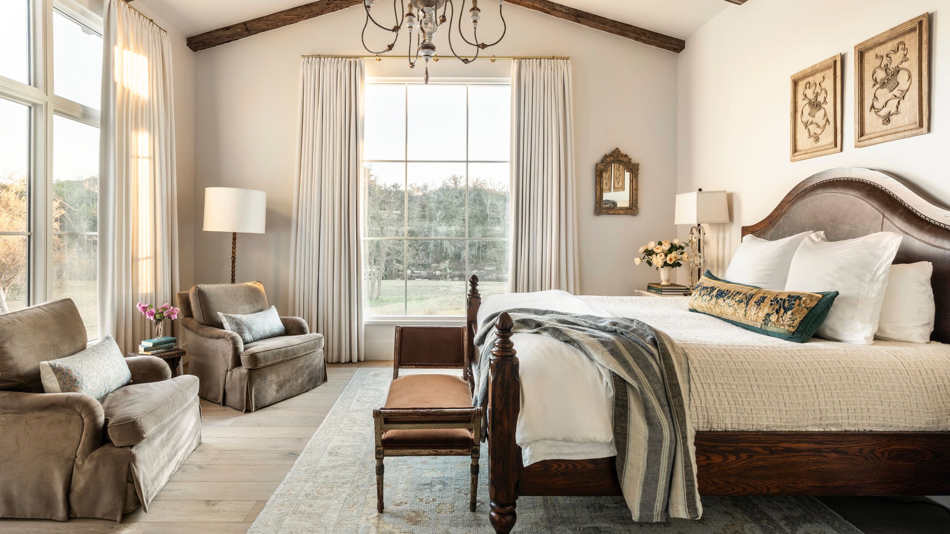 a layered farmhouse primary bedroom with neutral walls and rustic ceiling beams
