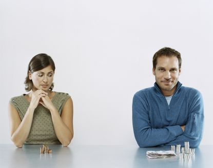 A man and woman of the same general age sit at a table. He has more coins in front of him than she does.