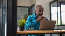 A man appears to be thinking while sitting at his desk in front of his laptop.