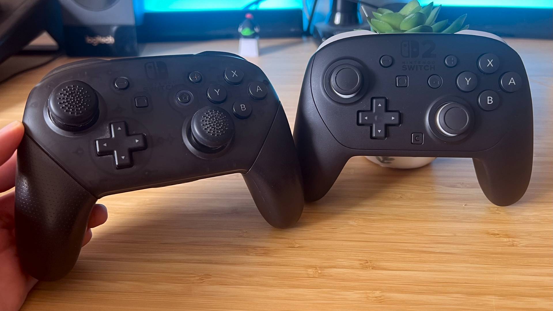 Original Switch 2 Pro Controller next to Switch 2 Pro Controller on woodgrain desk.