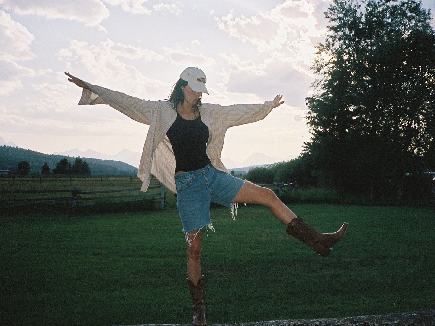 Kendall Jenner wears a beige button-down shirt, black tank, frayed-hem Bermuda denim shorts, and brown cowboy boots.