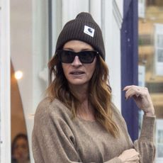 Julia Roberts wearing a brown sweater, dark brown pants, black flats, a navy bag, and brown knit hat
