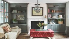 A modern living room with dark grey shelves, a white marble fireplace with a white panelled wall above, a red patterned ottoman, and a cream sofa with a patterned cushion.
