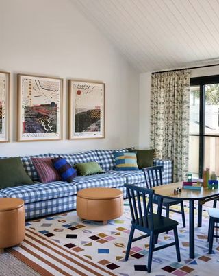 Bright living room with a geometric, multi-colored rug and a blue and white gingham sofa. There are two brown leather, round ottomans and a small navy wooden table in front of the sofa.