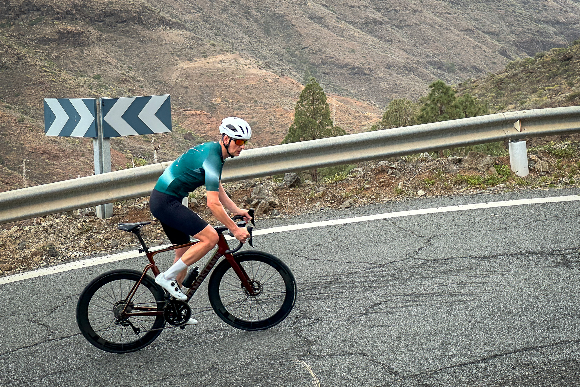 Man riding a bike out of the saddle wearing a white helmet, green jersey and black shorts, side on
