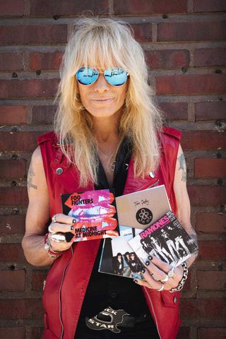 Michael Monroe in sunglasses, standing against a wall holding four CDs