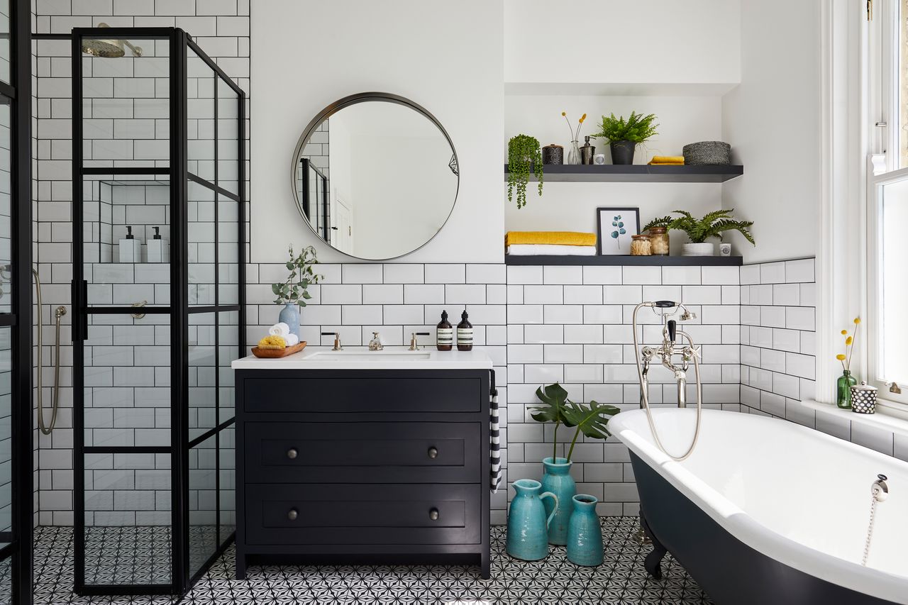 Clean white subway tiles in a monochrone bathroom with black shower panes, freestanding bath, shelving, sink vanity and geometric black and white flooring