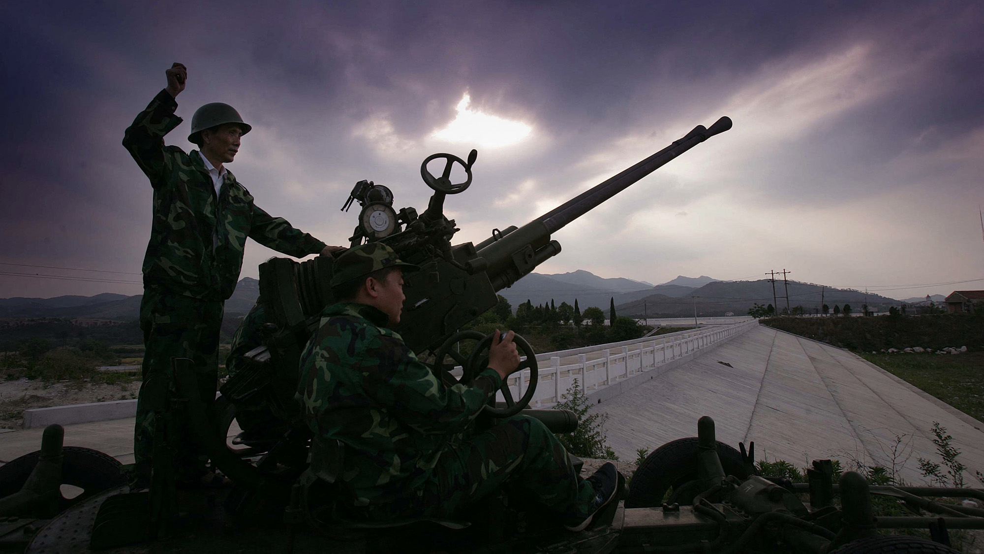 Two armed men wearing uniforms stand next to a small rocket launcher.