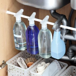 Closeup of interior of kitchen sink cupboard, plastic baskets containing cleaning products, plastic rod running across cupboard to hang bottles