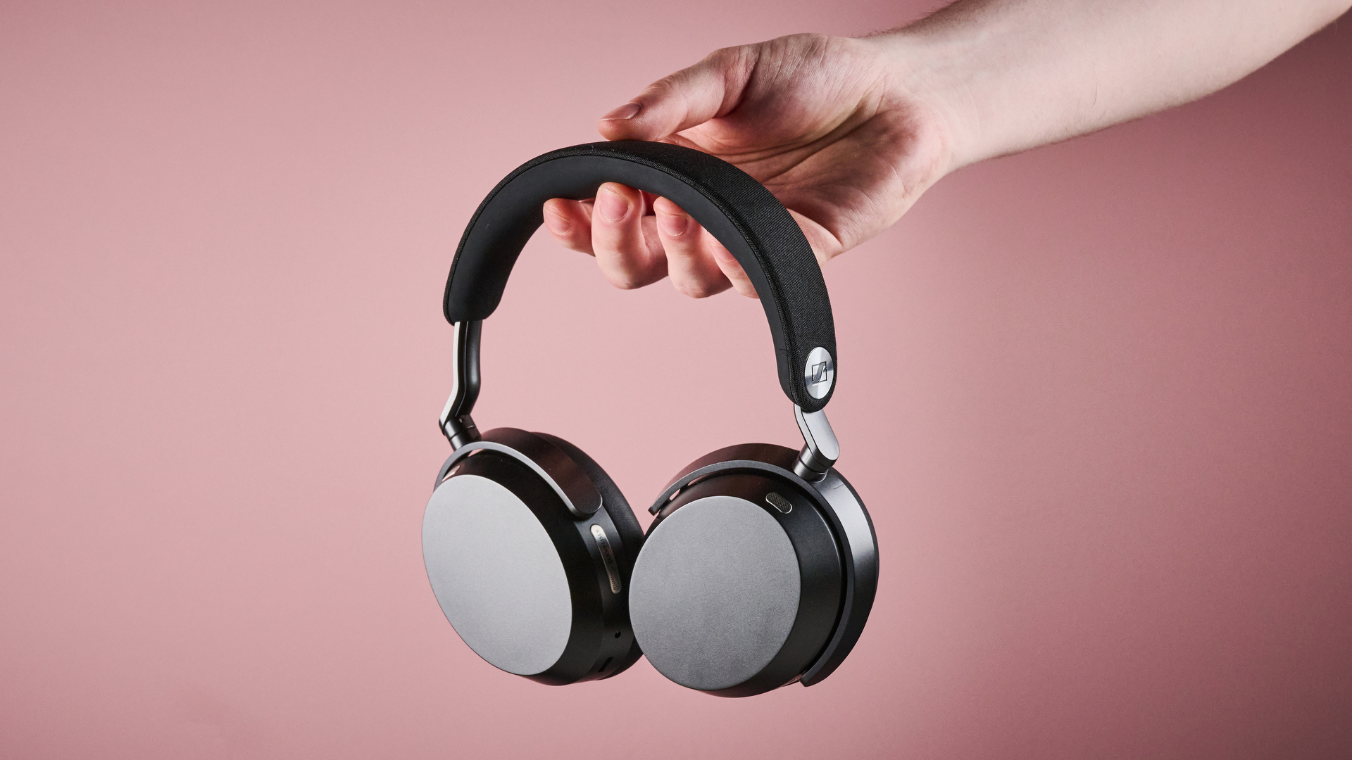A man's hand holds the Sennheiser Momentum 5 with its earcups folded flat in front of a pink background.