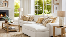 an inviting living room with a large white sofa with pillows in shades of beige and yellow on top, a small coffee table with decor on it, a printed rug, and a fireplace in the background on the left side