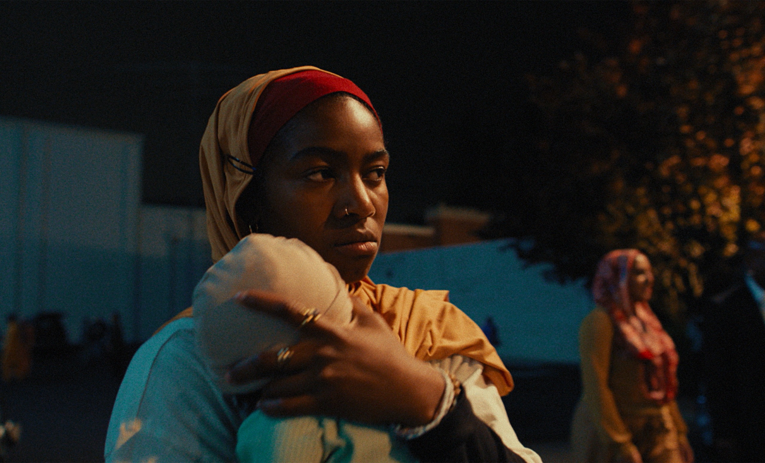 A teenage girl (Adia as Fatiha) wears a headscarf while holding a baby outdoors at night. Another woman is seen in the background. A still from the film 'Your Mother Your Mother Your Mother.'
