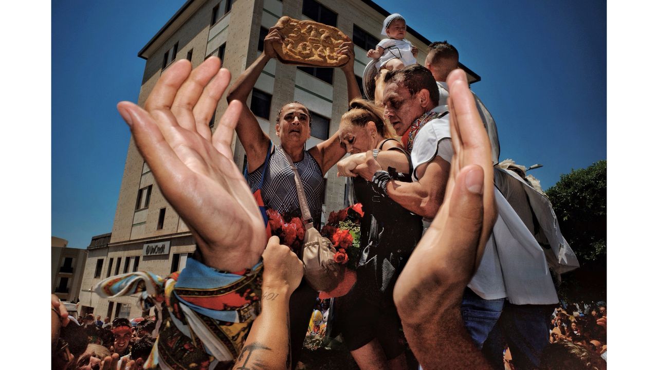 Crowd of people, including a man holding a flatbread, a woman with red flowers, and a baby, surrounded by raised hands under a clear blue sky