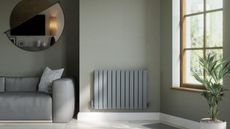 A light blur painted radiator mounted on a grey painted wall beside a lighter grey sofa. A half circle mirror hangs above the sofa. 