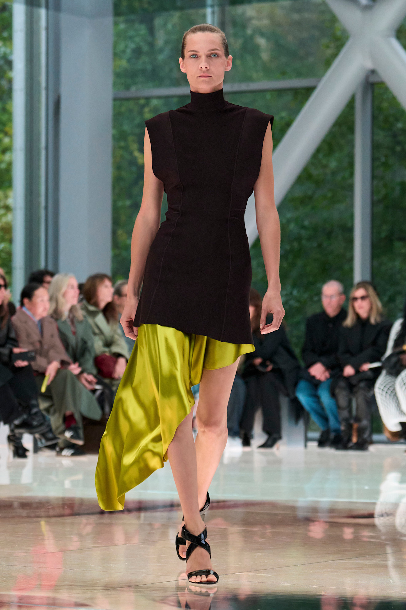 An Ala&iuml;a model wearing a high-neck cap sleeve blouse styled over a bright green asymmetrical slip skirt and black strappy sandals.