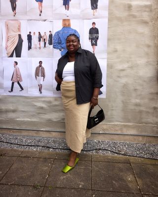 British plus size fashion influencer Abisola Omole poses on a sidewalk in Copenhagen wearing an open black button-down shirt, white top, tan midi skirt, black shoulder bag, and green Prada square-toe mules