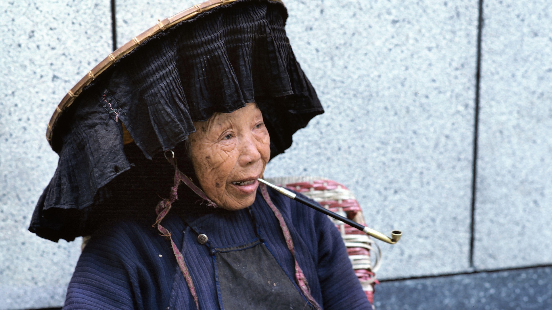 An older woman balances a long, thin pipe in her mouth. She wears a large, circular hat with dark fabric hanging down around the rim.