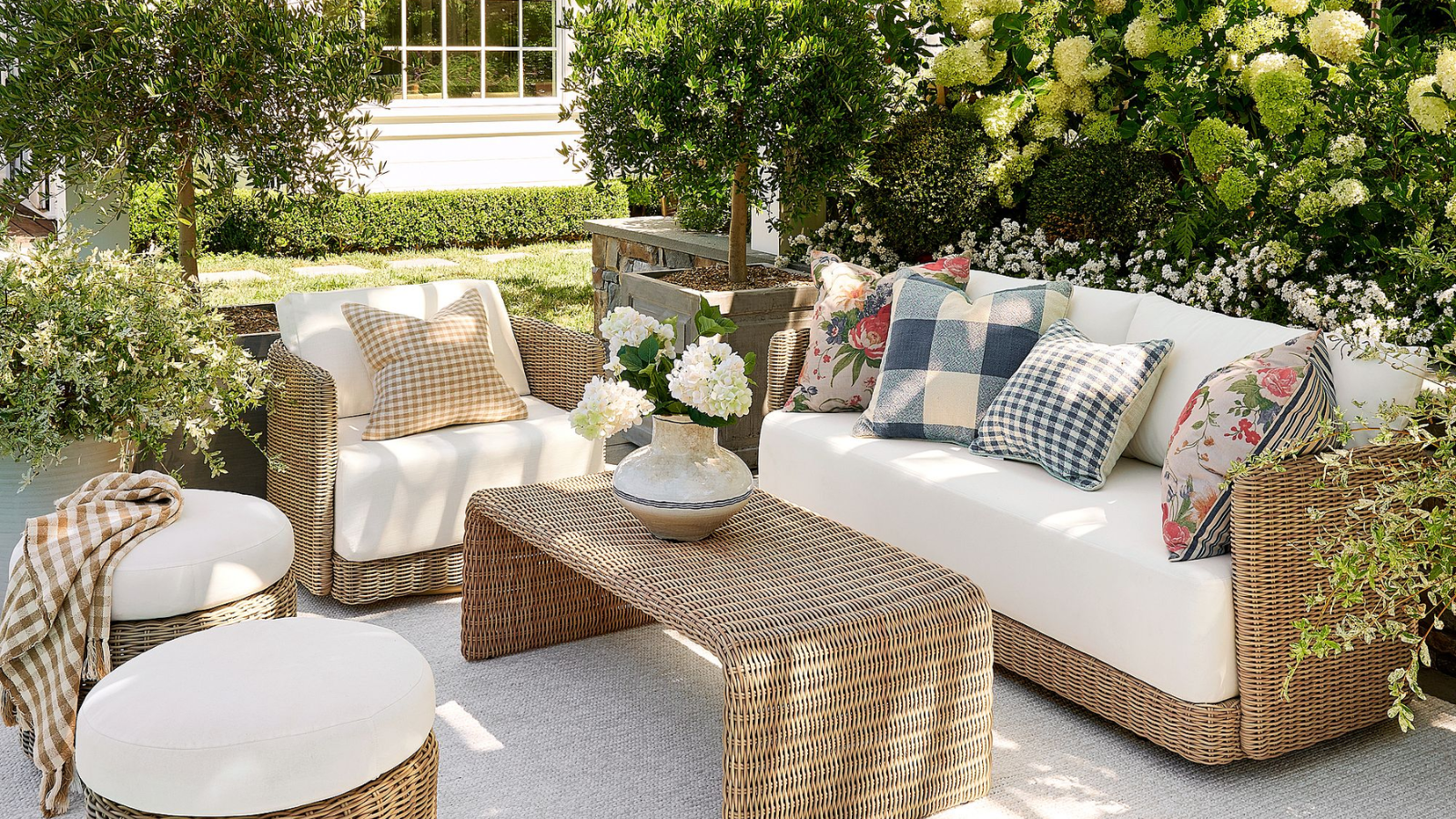 an outdoor living room with wicker furniture, white cushions, colorful pillows, a coffee table, a vase of flowers, two ottomans, and lots of greenery around the scene