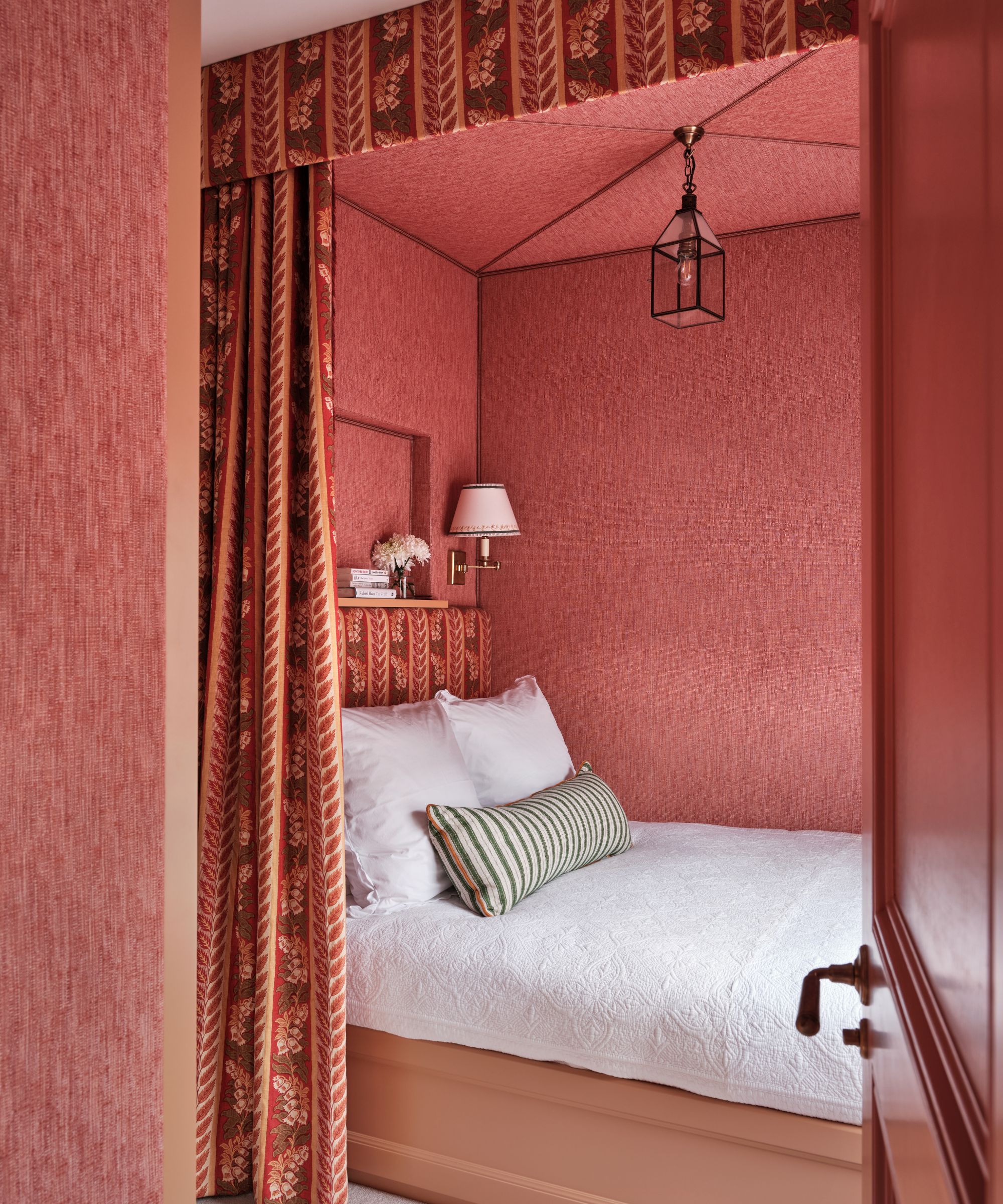 Bed nook with coral grasscloth walls, a floral red pelmet curtain, a matching upholstered headboard, white linen bedding with a striped green bolster cushion, and a glass lantern hanging above.
