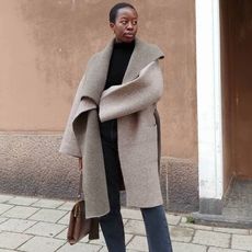 European fashion influencer Sylvie Mus poses on a Paris sidewalk with an oversize neutral coat, black mock neck top, vintage top-handle bag, jeans, and a brown bag