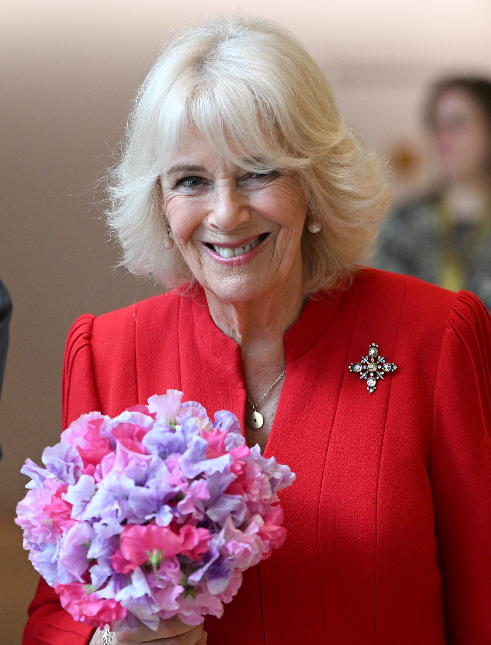 Queen Camilla holding flowers and wearing a red dress with a diamond brooch