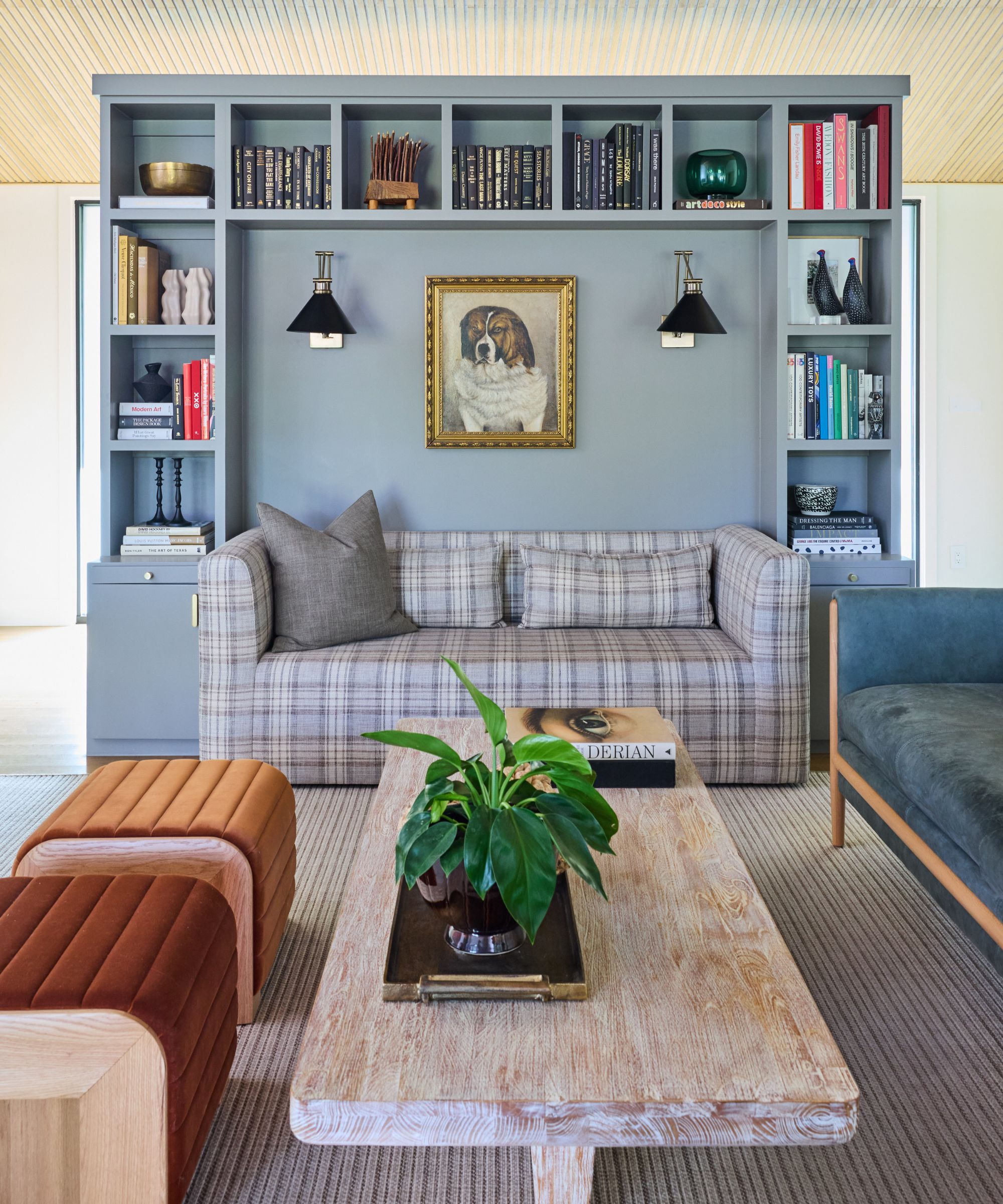 A living room with a gray bookshelf in the middle of the room, a plaid gray sofa, a rustic coffee table, a blue sofa, and brick-colored seating.