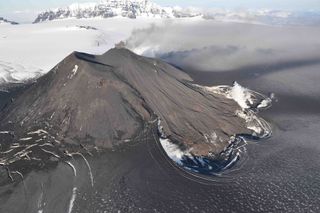 Aerial view of the erupting intracaldera cone at Veniaminof, Aug. 18, 2013. This photo shows new lava flows produced since the eruption started on June 13. These flows, roughly 3000 feet (900 meters) long, have descended the southwest flank of the cone and traveled onto the ash-covered snow and icefield. Melting of the snow and ice has created the depressions beneath the young flows.