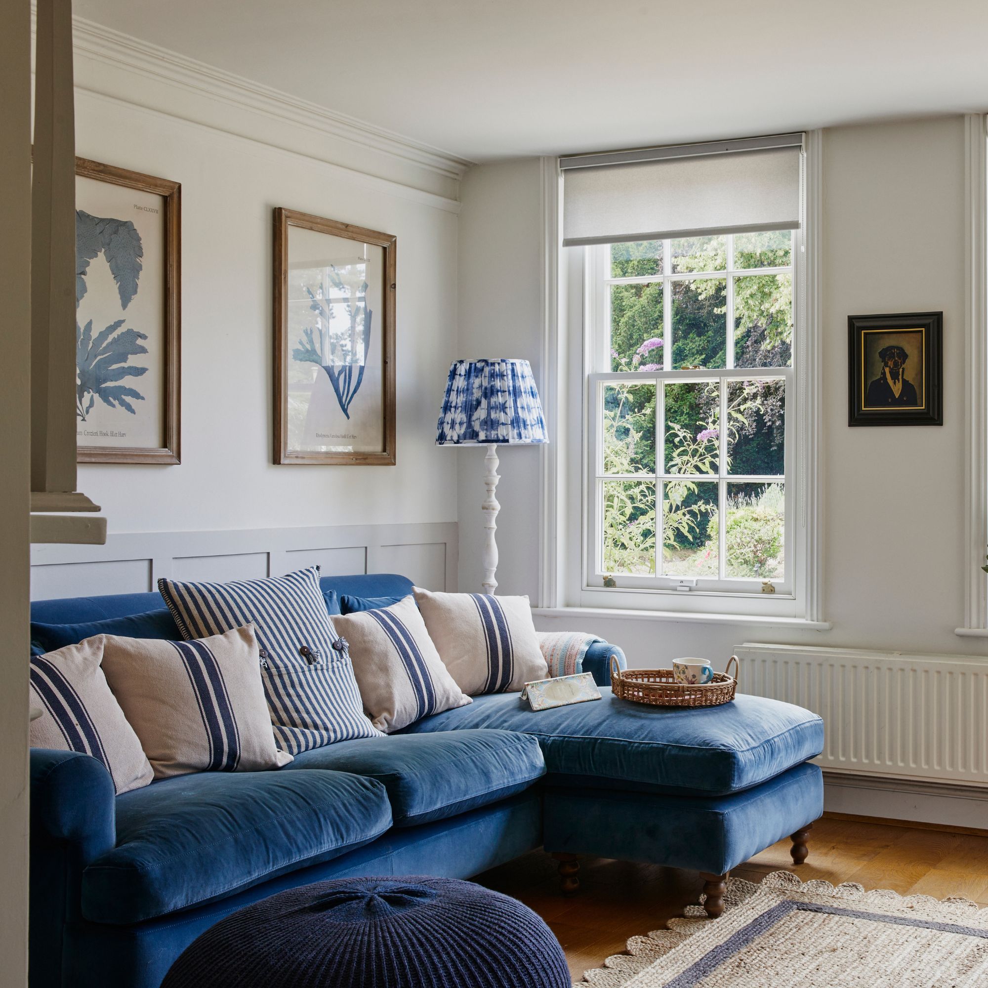 Light-filled living room with artwork on the walls, a blue velvet sofa, blue and white floor lamp, navy footstool and sash windows