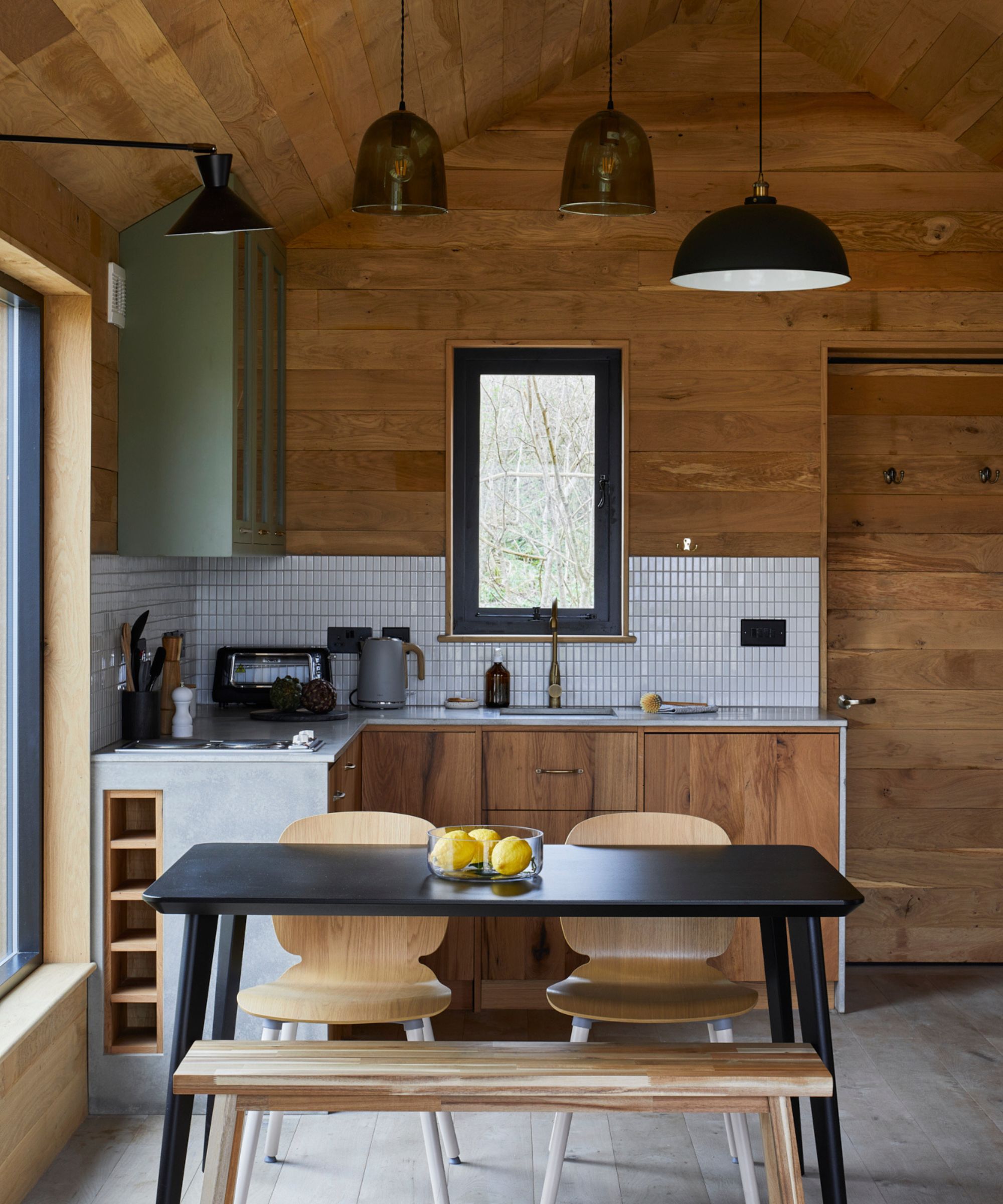 A modern treehouse kitchen with vaulted timber ceilings and mixed wood finishes. The space features a black dining table with natural wood chairs and bench seating, alongside flat-panel cabinetry and a tiled splashback.