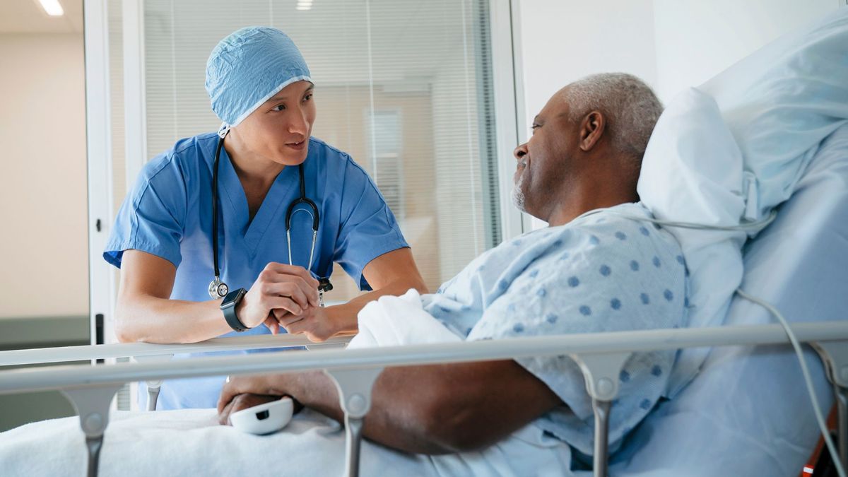 surgeon wearing blue scrubs speaks to a black male patient resting in a hospital bed. both are smiling