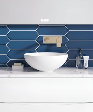 A white vessel sink on top of a white vanity unit, with a dark blue splashback with a gold faucet behind it