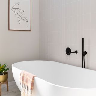 A modern white ceramic bath with decorative multicoloured bath towel, matt black shower accessories fixed to wall, taupe vertical bathroom tile decor on walls and framed wall art,