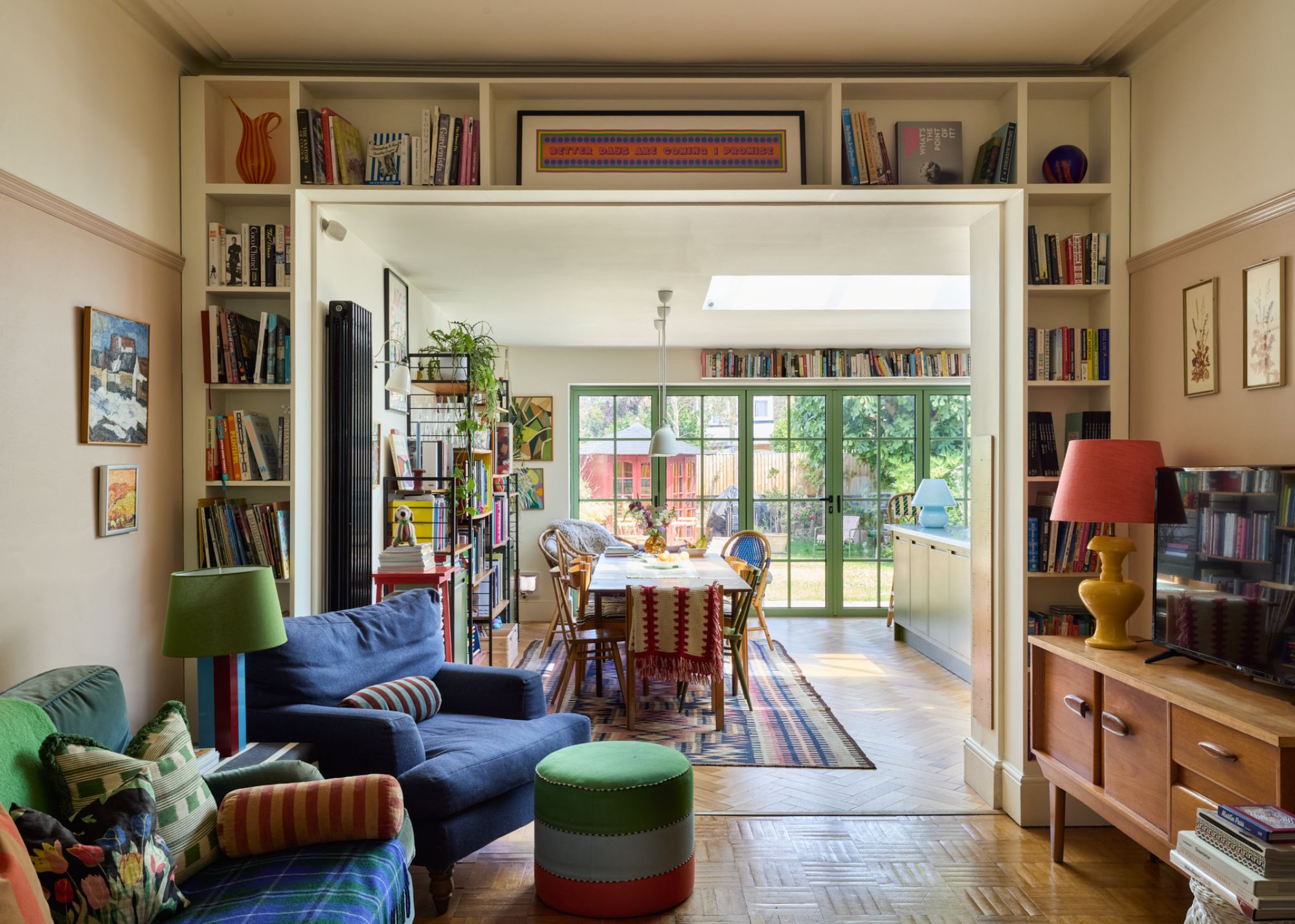 A living room leading into an open-plan kitchen and dining room. Wooden flooring, open shelving built into the doorway, two dark blue armchairs with a green lamp and footstool, and a vintage midcentury wood sideboard.